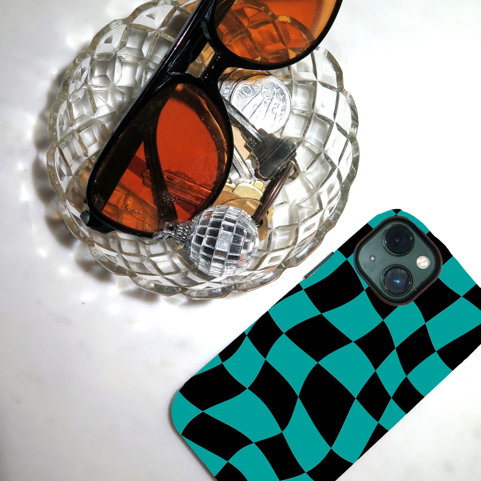 Orange sunglasses on a clear glass dish with a checkered phone case next to it.